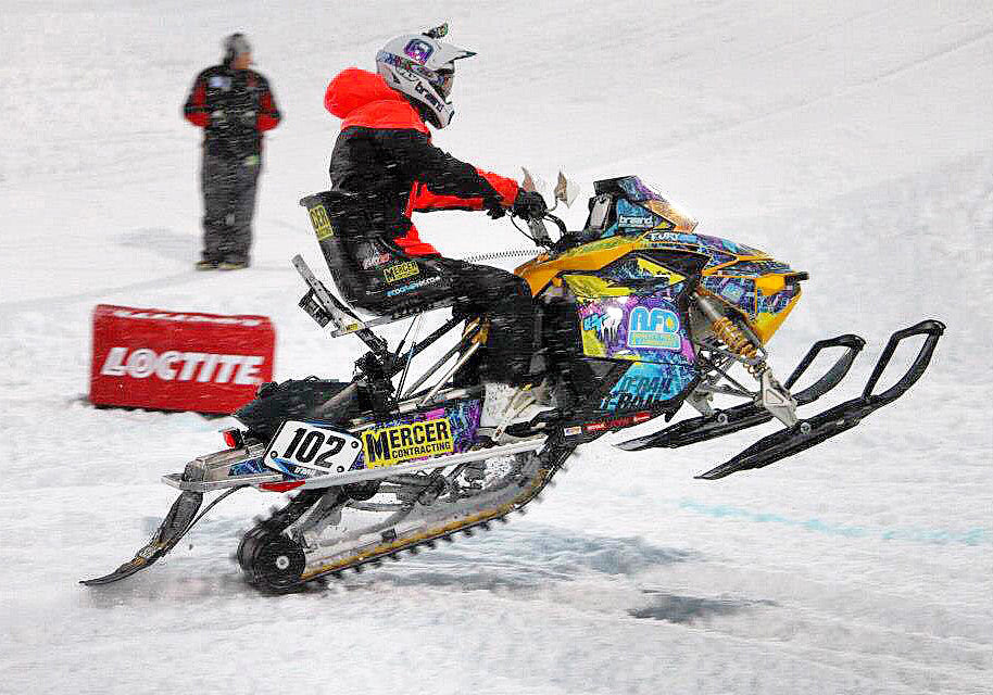 Darryl Tait riding a snow sled pulling a wheelie, wearing a helmet and bright jacket