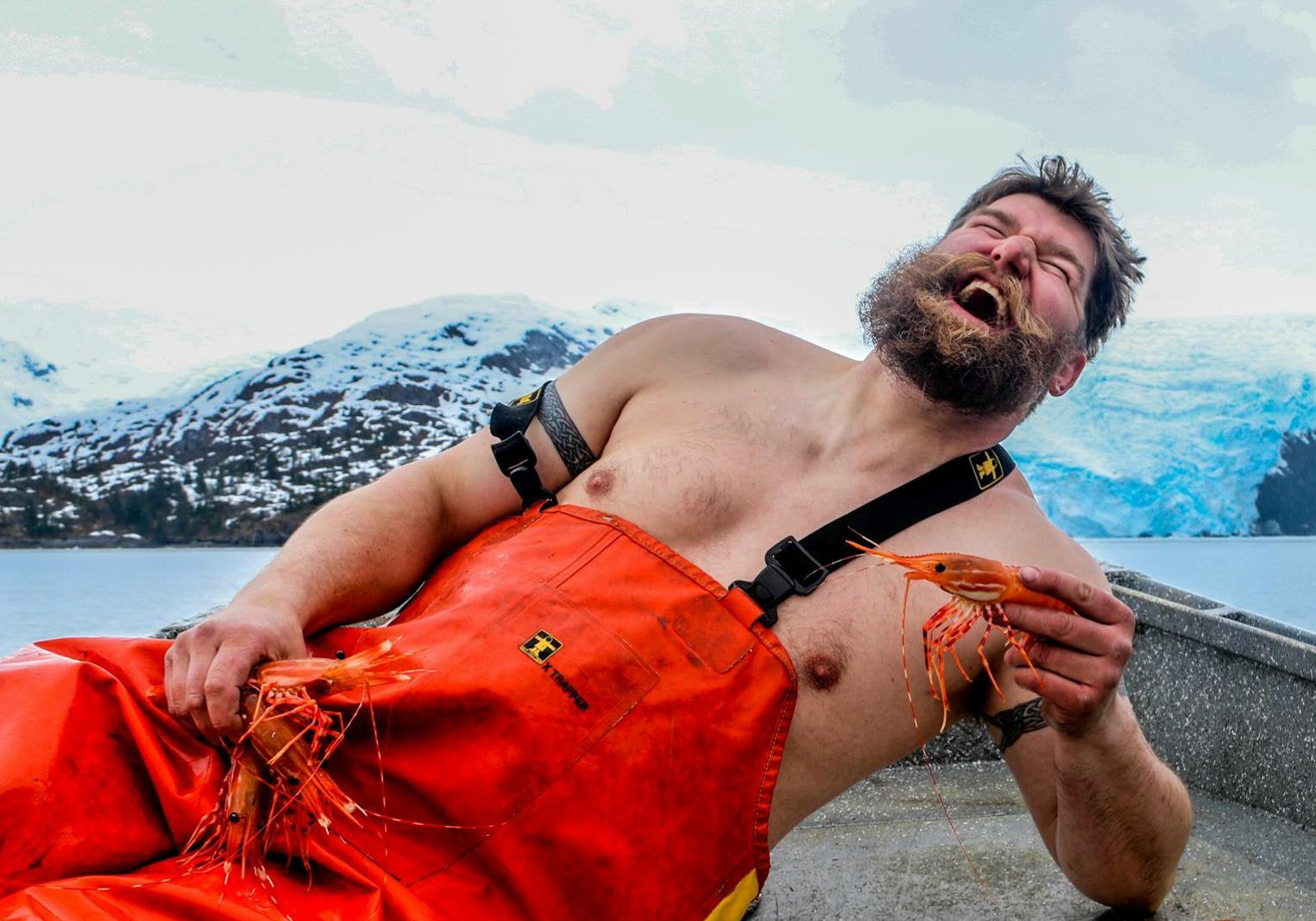 Ira Edwards, wearing bright orange overalls, holidng prawns while sitting and leanign back on a boat with Alaskan mountains and sea in the background and grinning wildly. 