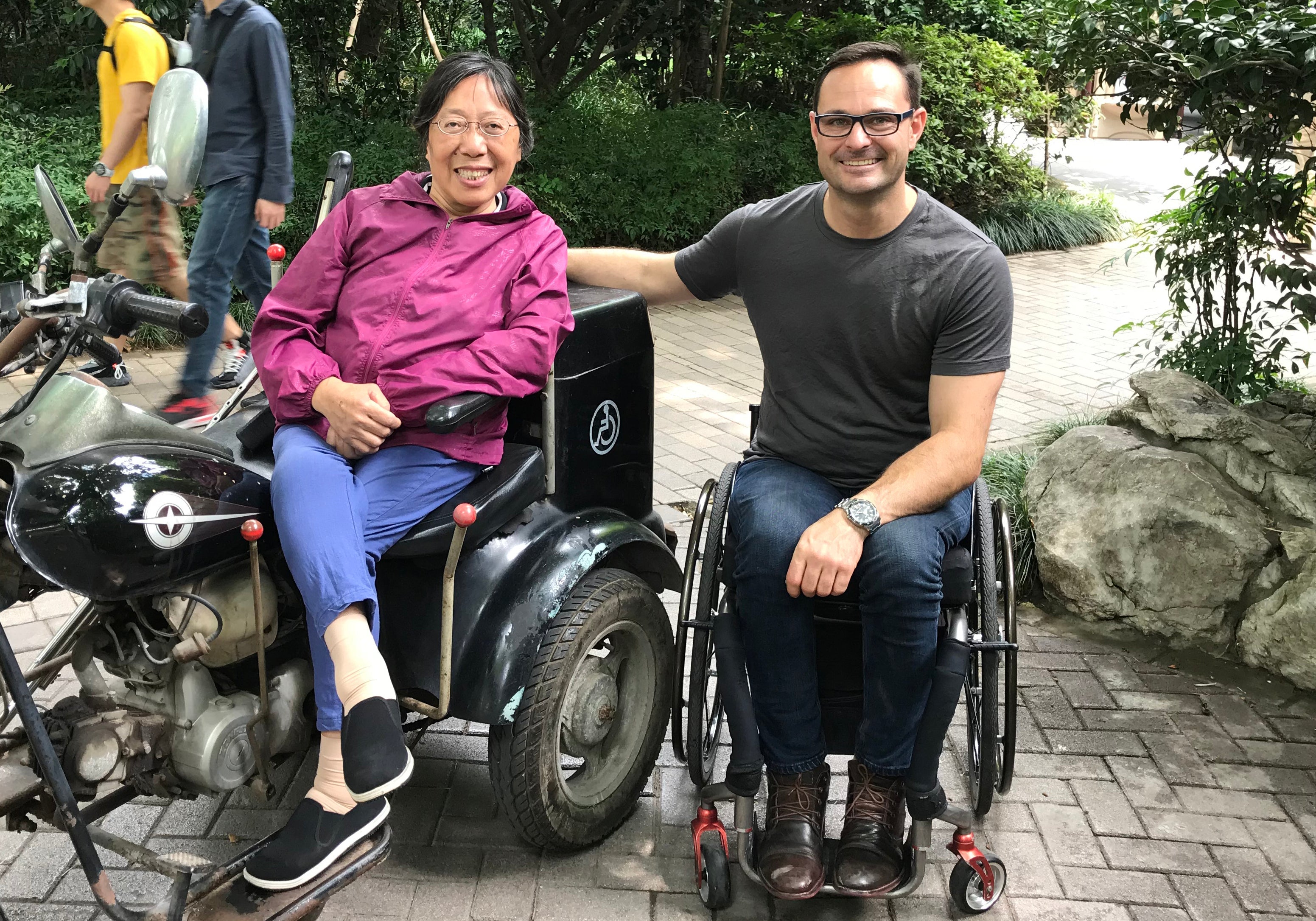Two adults are sitting side by side outdoors on a paved path; one sits on a three-wheeled motorized trike-style mobility vehicle, and the other sits in a manual wheelchair. They are smiling, relaxed, and surrounded by greenery.