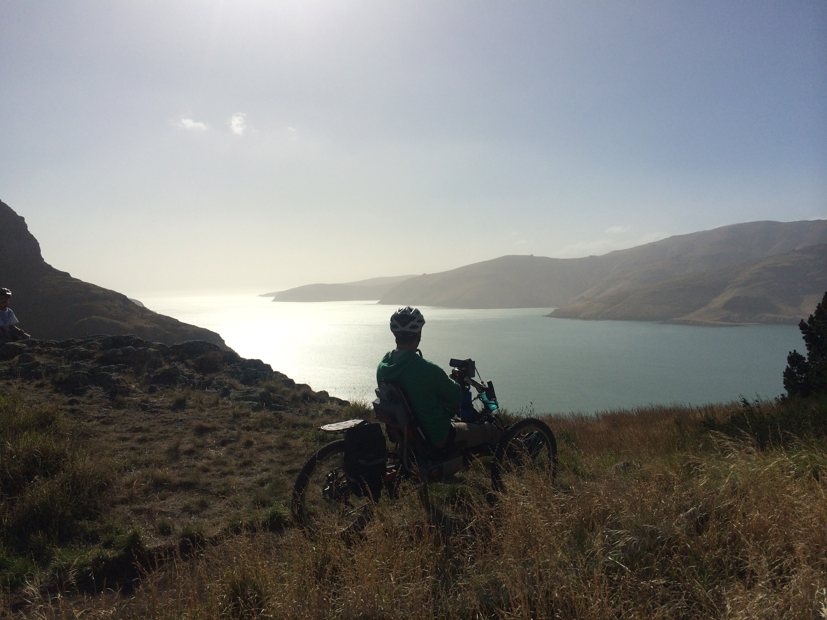 A picture of Mike sitting on a hand-cycle with golden tussock all around and looking over Lyttelton harbour with blue sky and sun shining.