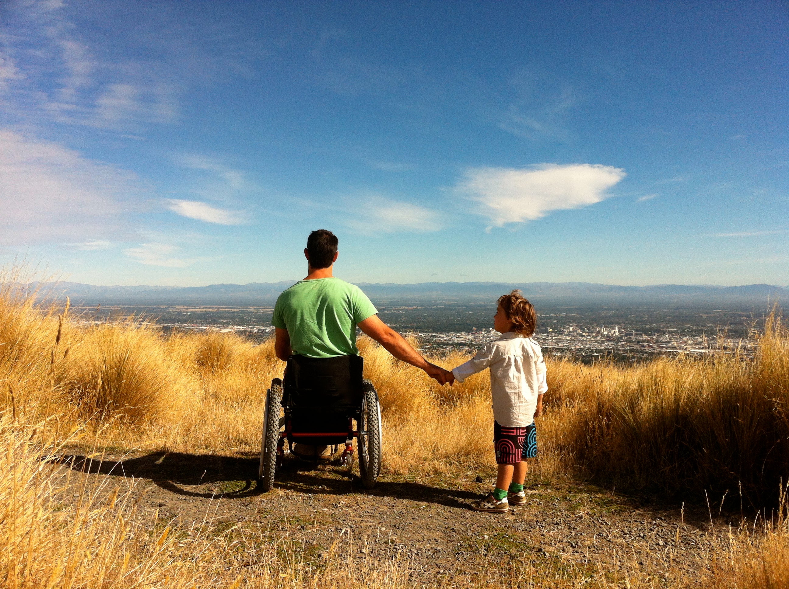 Mike in wheelchair holiding hands with son looking out from the port hills.