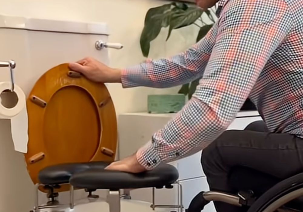 A man in a wheelchair adjusts a raised, padded toilet seat attachment in a bathroom. He is wearing glasses and a checked shirt, with the wooden toilet lid lifted. The setup helps make the toilet more accessible for easier transfers.