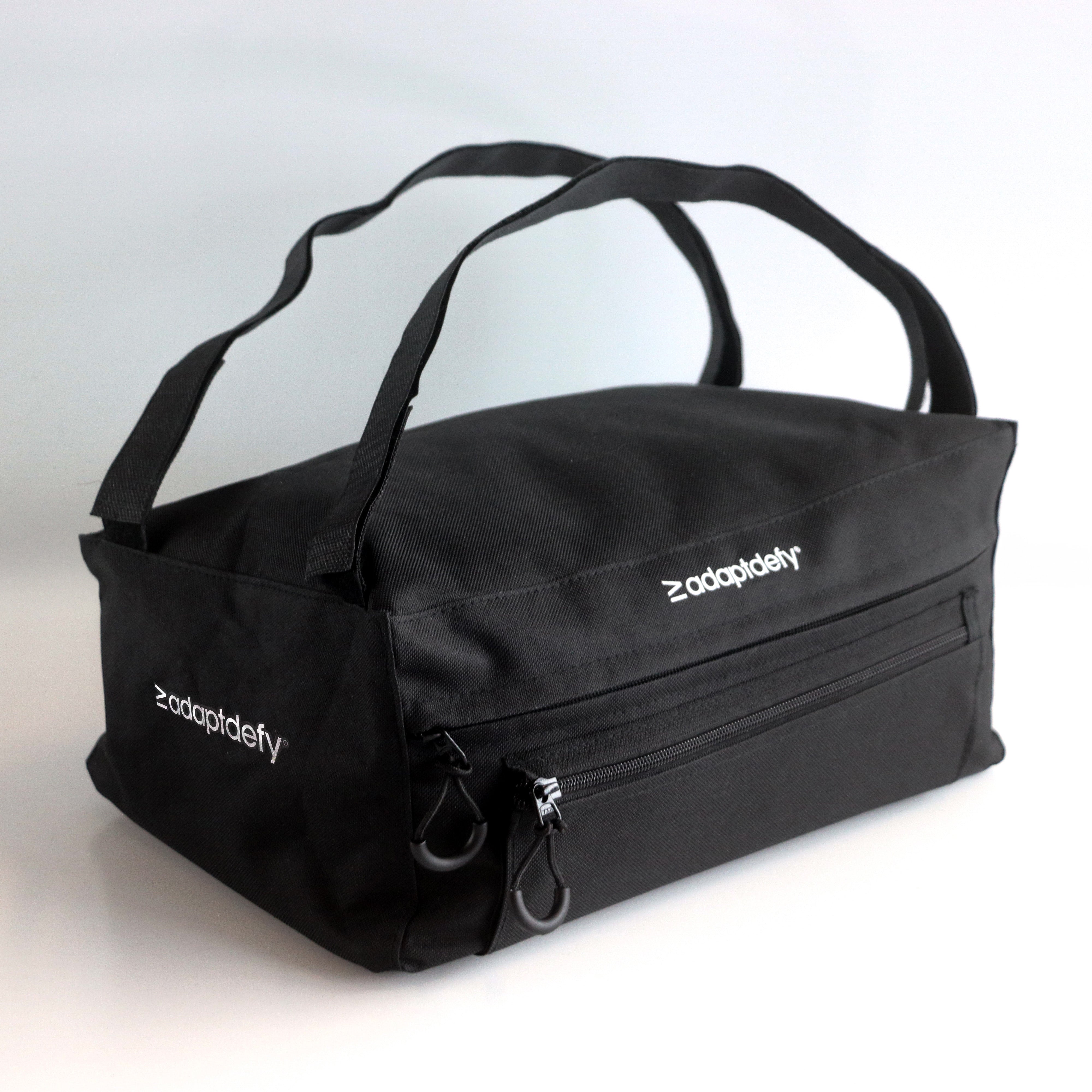 Black wheelchair bag from 45 degree sideways angle. Details show, adjustable velcro straps, zipper loops on the front, and a main pocket. "Adaptdefy" branding on both sides and front 