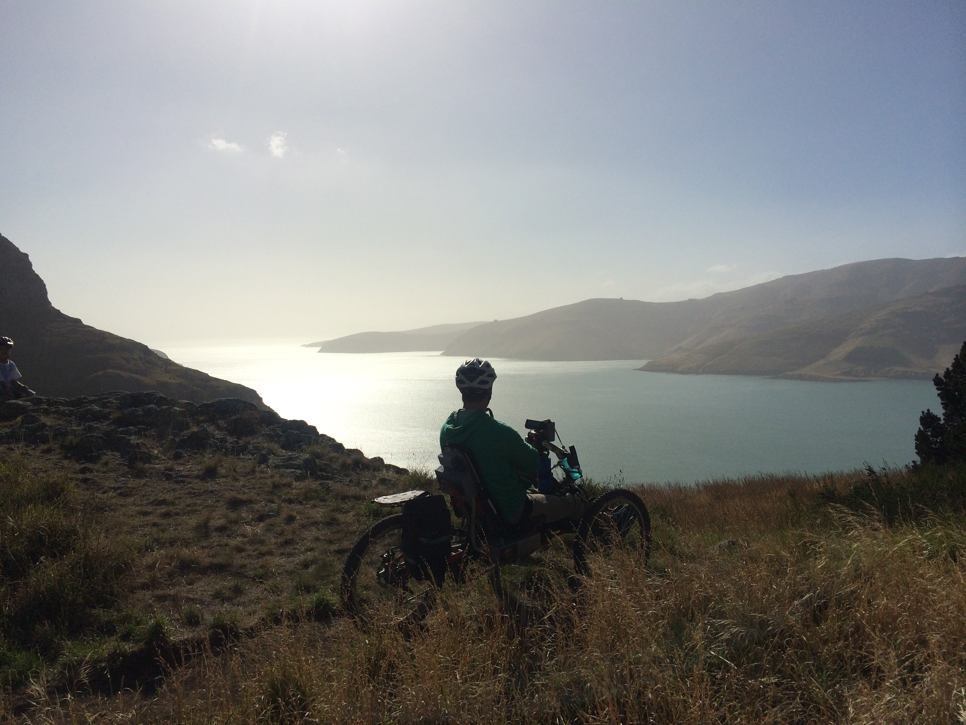 A picture of Mike sitting on a hand-cycle with golden tussock all around and looking over Lyttelton harbour with blue sky and sun shining. 