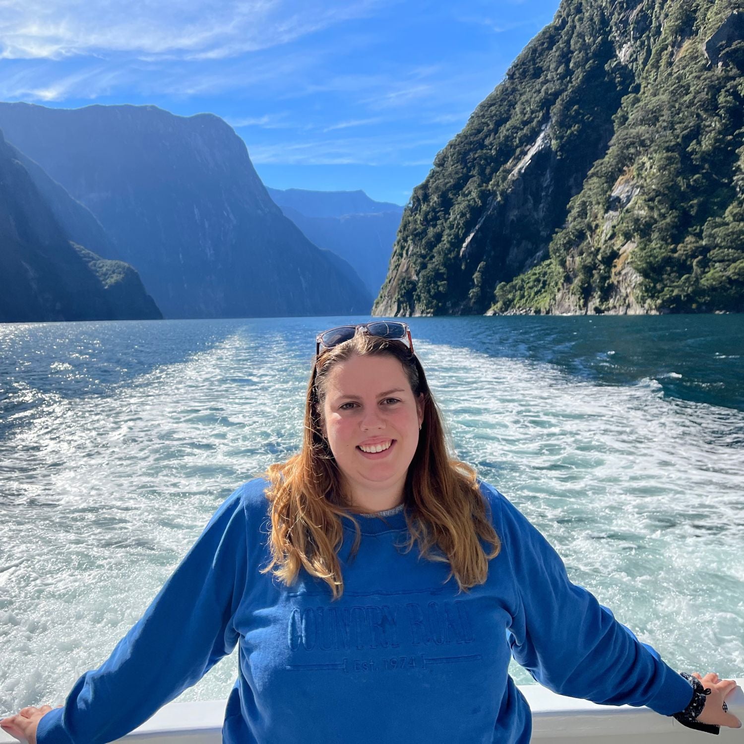 Woman in a blue sweater standing on a boat with a scenic mountainous landscape in the background