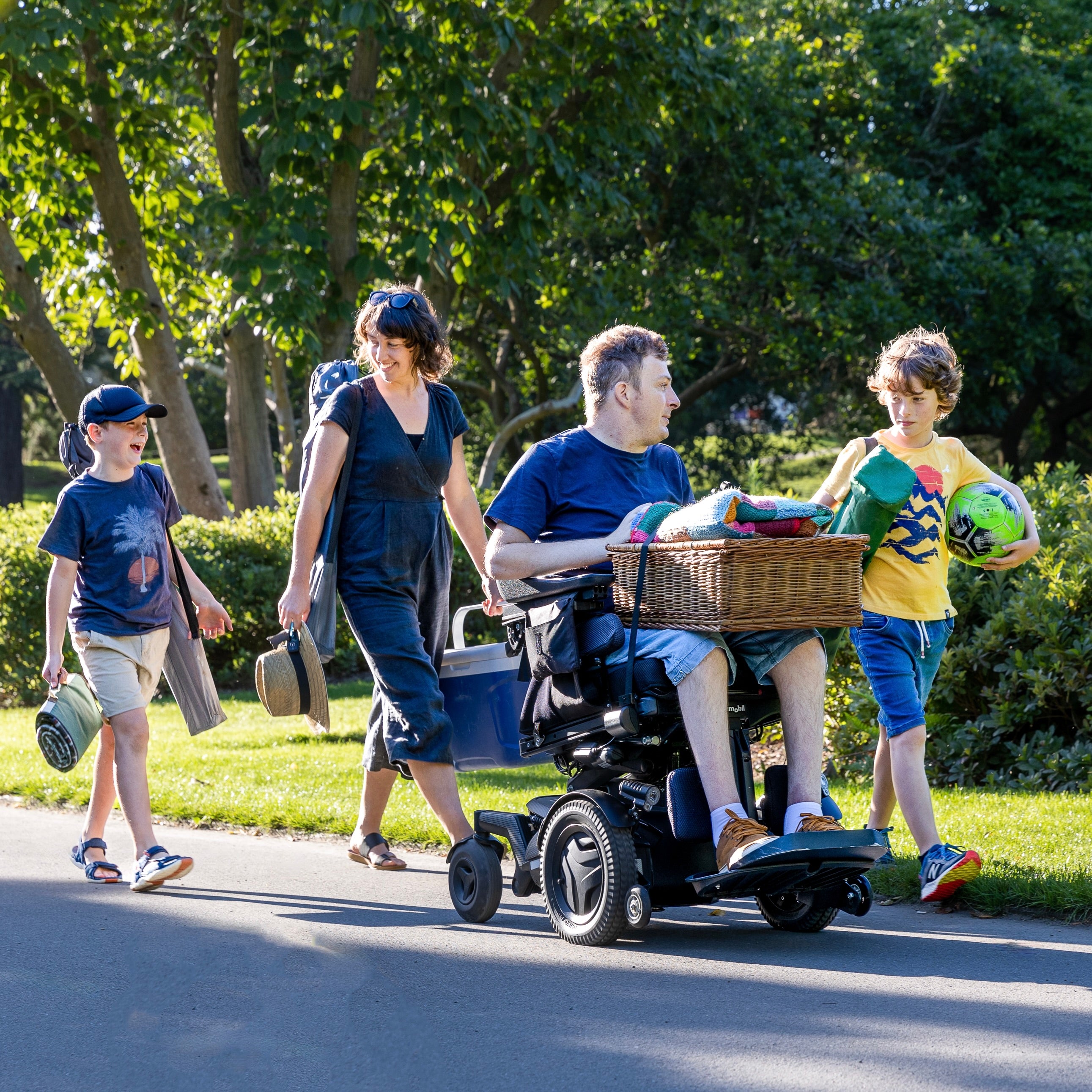 Man in a power wheelchair at the park with his family. He is carrying a picnic box and blanket on his lap which is secured with a LapStacker XD. He is smiling as he confidently wheels along. 