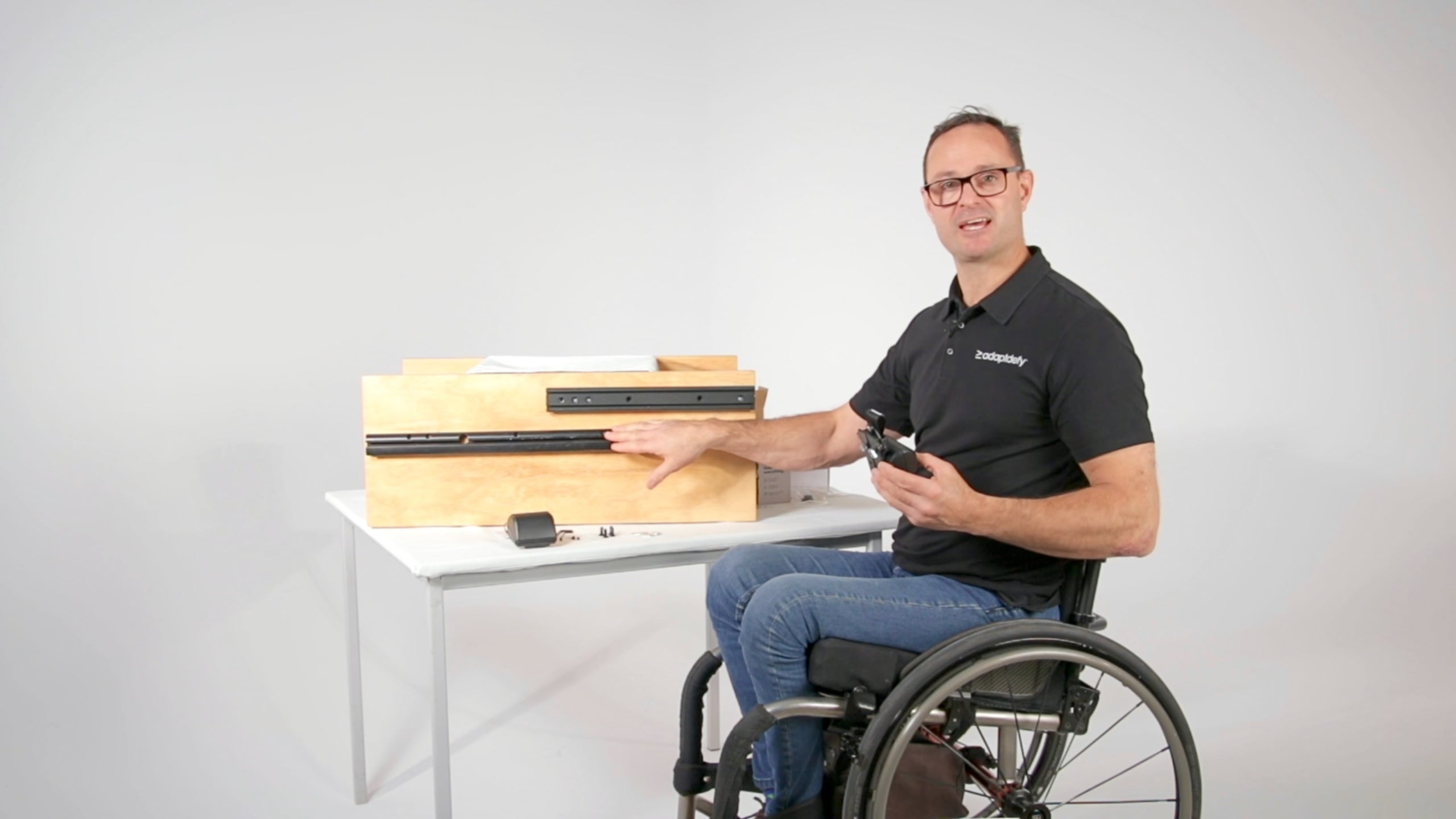 A man in a wheelchair shows what's in the box and how to install a LapStacker XD on the T-slot of a power wheelchair. He shows how the mounting accessories are compatible with power wheelchair brands including Amylior, iBOT, Invacare, Merits, Ottobock, Permobil, Quantum, and Quickie.