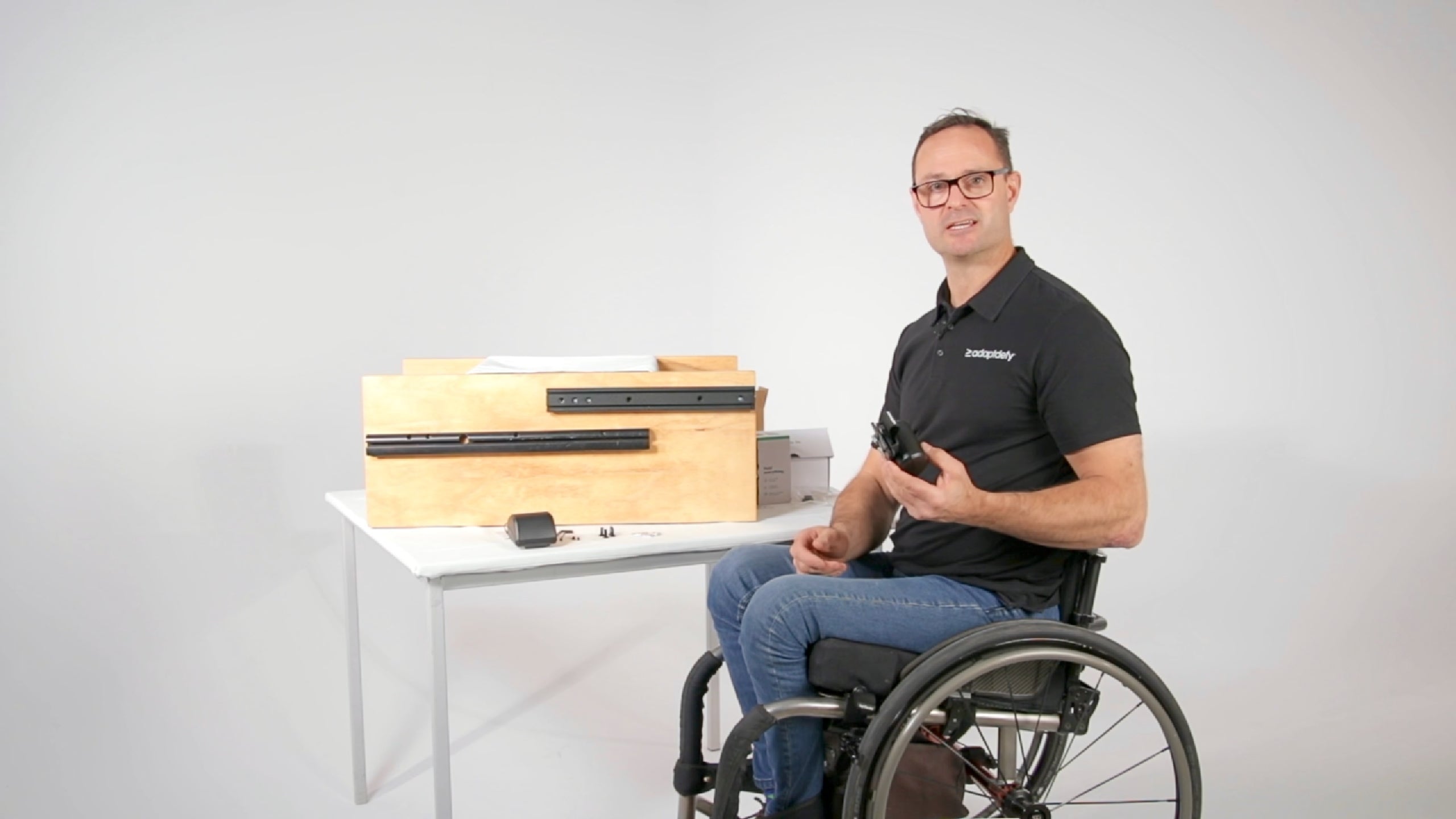 A man in a wheelchair shows what's in the box and how to install a LapStacker XD on the T-slot of a power wheelchair. He shows how the mounting accessories are compatible with power wheelchair brands including Amylior, iBOT, Invacare, Merits, Ottobock, Permobil, Quantum, and Quickie.
