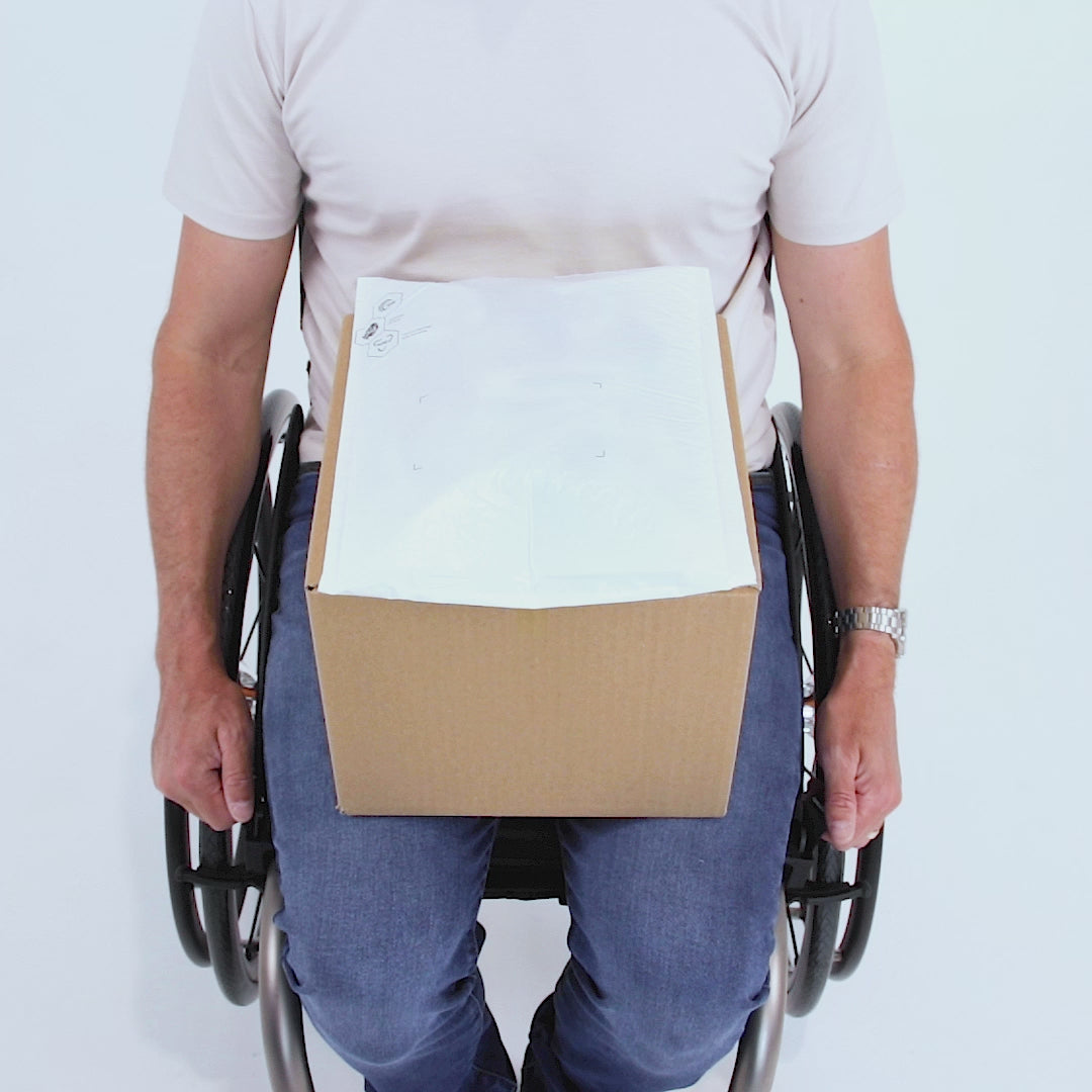 Man in a manual wheelchair with a white background, using LapStacker to secure a range of different items, one after the other. Items include, boxes, laundry, shopping, luggage, laptop, eggs, pizza, basketball etc...