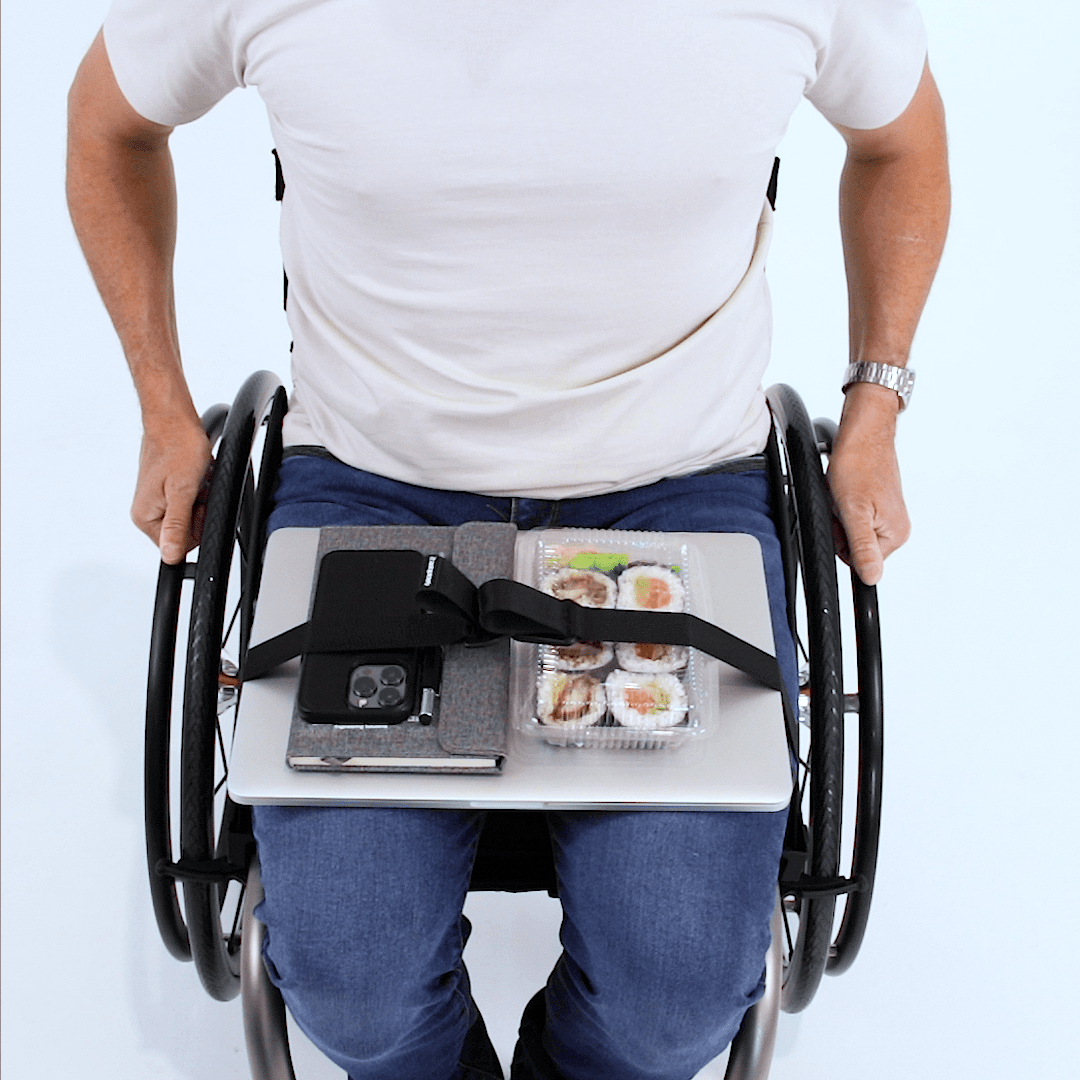 Overhead view of a man seated in a manual wheelchair with a laptop, notebook, phone and packet of sushi secured with LapStacker Flex